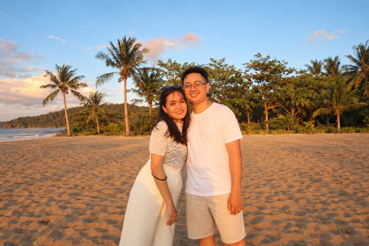 Carlo and Kristinne on a beach at sunset, both in white, palm trees behind them.
