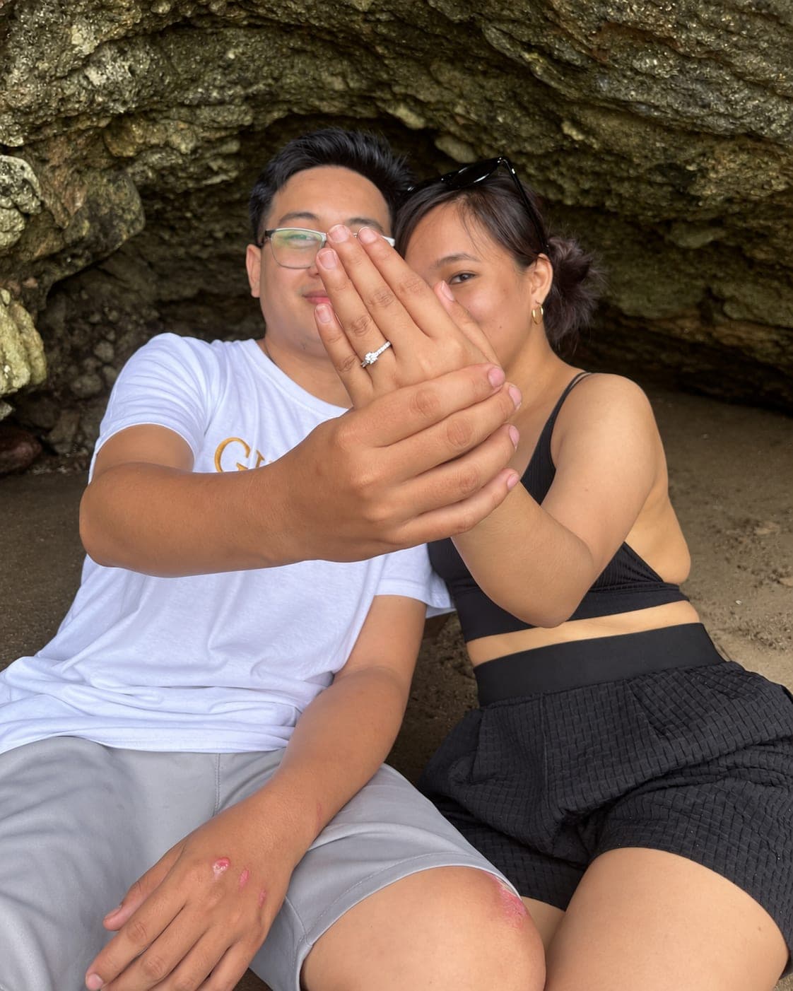 Kristinne holding up her hand to show her engagement ring, Carlo beside her in a rock cave by the sea.