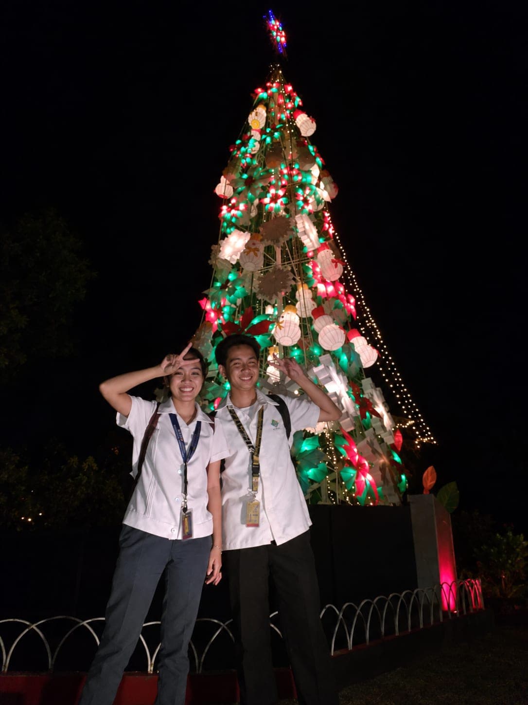 Carlo and Kristinne in school uniforms making peace signs in front of a lit Christmas tree at night.
