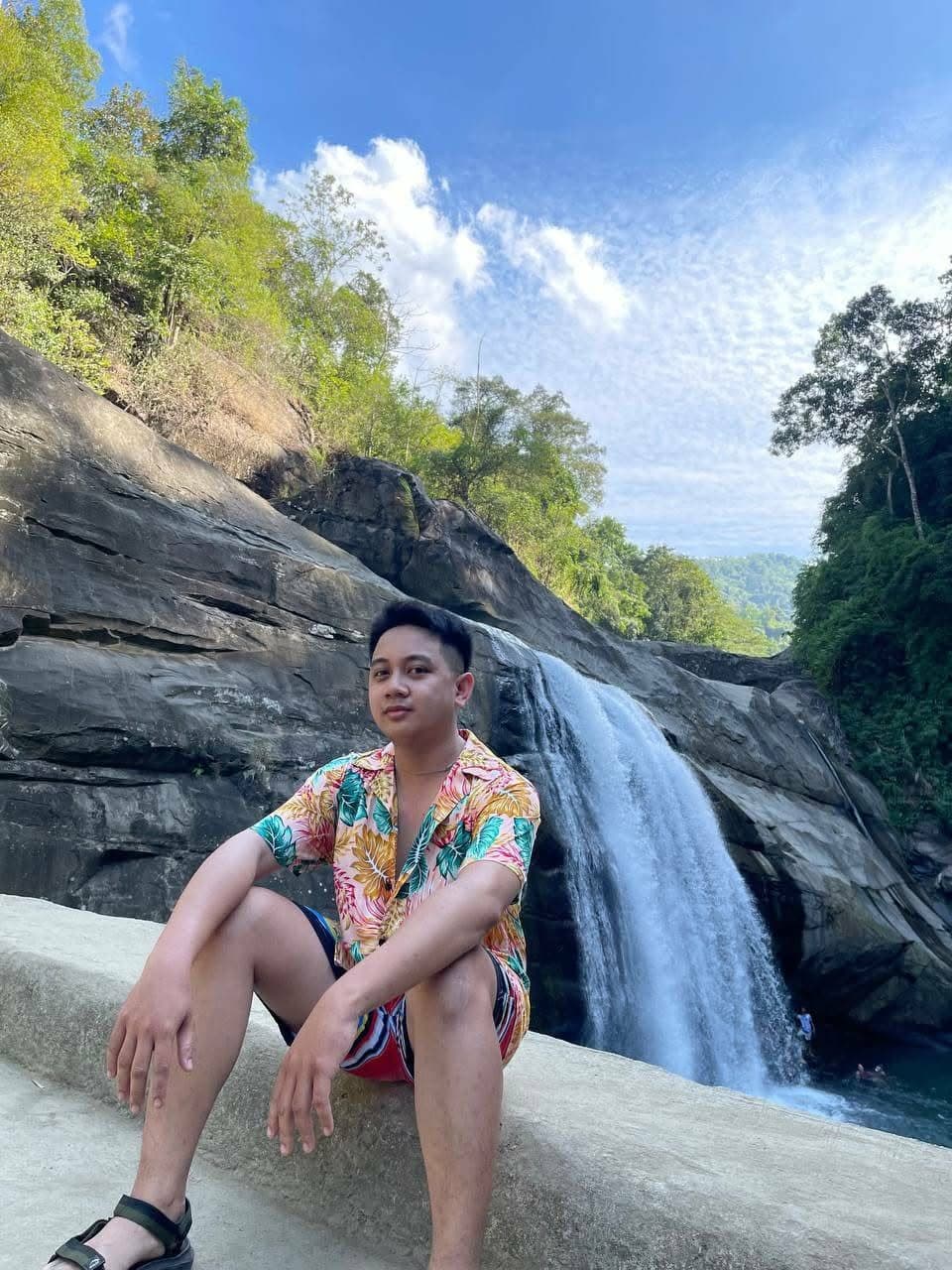 Carlo sitting on a rock in front of a waterfall, in a colourful shirt.